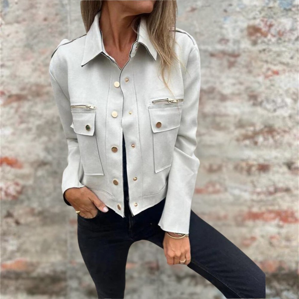 Donna con giacca bianca elegante, tasche frontali e bottoni dorati, posa contro muro di mattoni. Moda autunnale, stile casual chic.