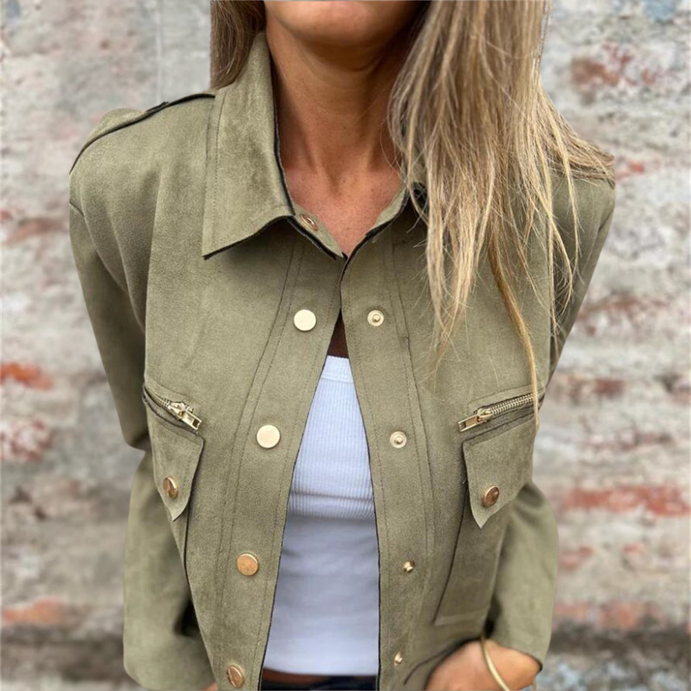 Donna con giacca verde militare, bottoni dorati, tasche con zip, su sfondo di muro in mattoni. Moda autunnale, stile casual chic.