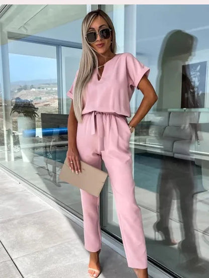 Donna con occhiali da sole indossa elegante tuta rosa, tiene una borsa beige. Sfondo moderno con vetrate. Moda femminile, abbigliamento estivo.