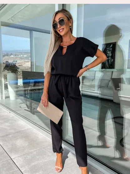 Donna elegante in tuta nera, occhiali da sole, sandali bianchi, con clutch beige, in terrazza moderna con vista panoramica. Moda estiva chic.