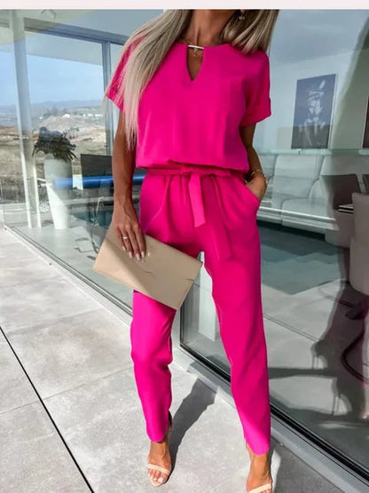Donna in tuta fucsia elegante, con pochette beige, in ambiente moderno con vetrate. Look estivo chic, moda femminile contemporanea.