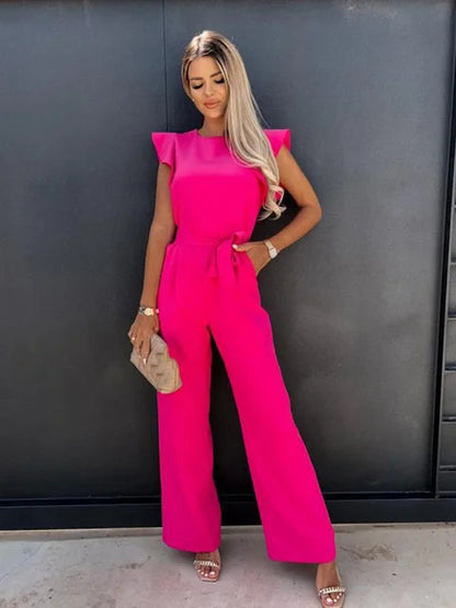 Donna in tuta rosa fucsia elegante, posa contro parete nera. Moda femminile, abbigliamento estivo, stile chic. Sandali beige e borsa a mano.