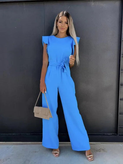 Donna in tuta blu elegante, con cintura in vita, posa davanti a parete nera. Indossa sandali con tacco e borsa beige. Moda femminile estiva.