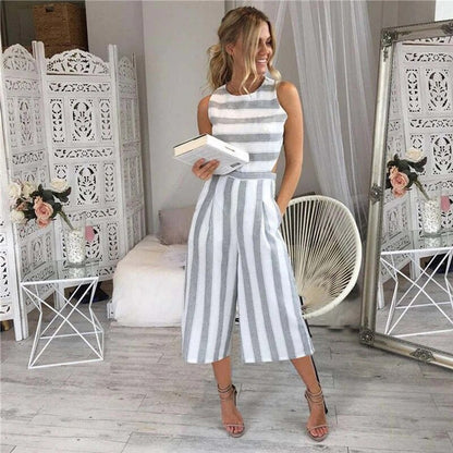 Donna in tuta a righe bianche e grigie, con libro in mano, in salotto elegante con decorazioni floreali e mobili bianchi. Moda estiva chic.