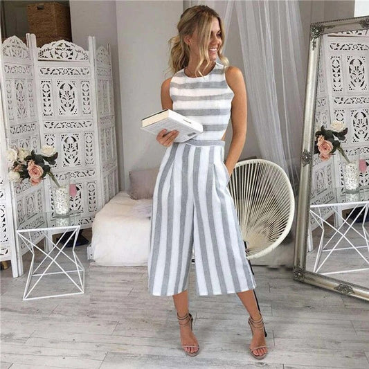 Donna in tuta a righe bianche e grigie, con libro in mano, in salotto elegante con decorazioni floreali e specchio. Moda estiva, abbigliamento casual.