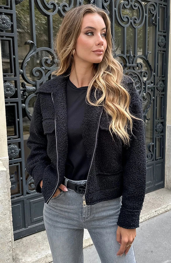 Donna con giacca nera in lana, maglia nera e jeans chiari, posa davanti a un cancello decorato. Moda autunnale, stile casual chic.