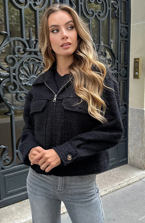 Donna con giacca nera in lana, jeans grigi, davanti a un portone decorato. Moda autunnale, look casual chic, capelli ondulati.