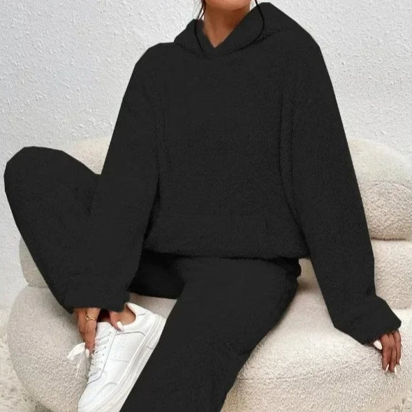 Donna seduta su poltrona indossa tuta nera invernale con cappuccio, pantaloni larghi e sneakers bianche. Look casual e comodo per l'autunno.