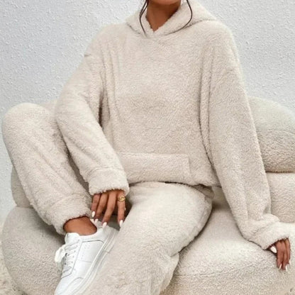 Donna seduta su poltrona indossa tuta in pile beige, comoda e calda, con cappuccio. Moda casual autunnale, abbigliamento loungewear trendy.