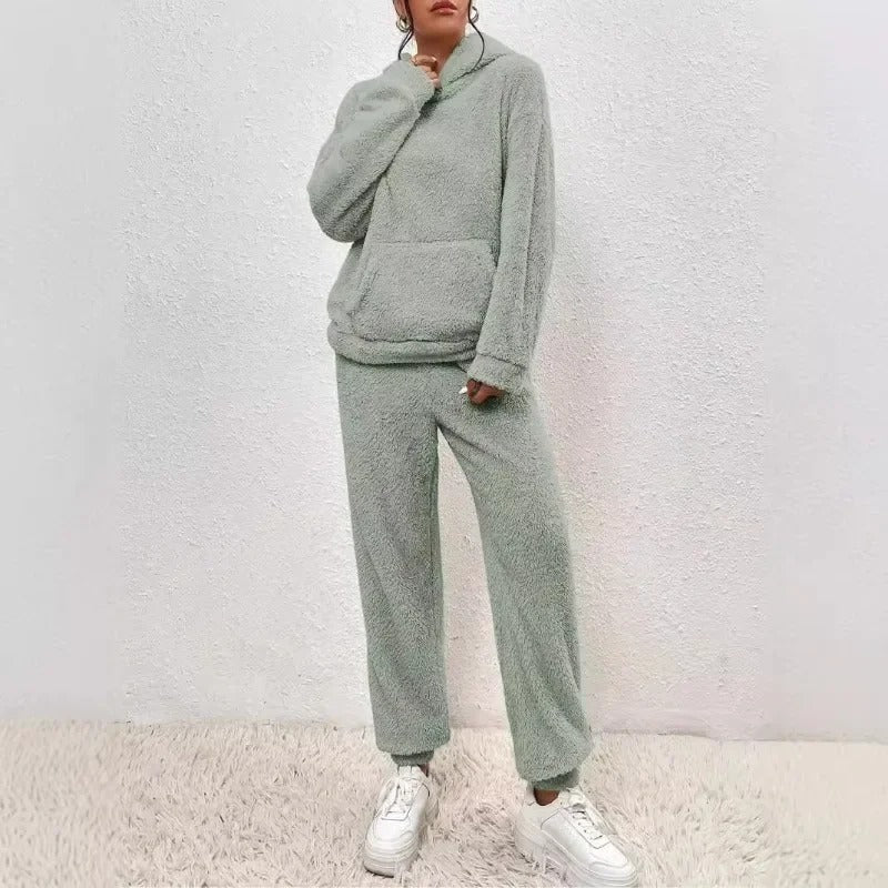 Donna in tuta felpata grigia, con cappuccio e tasca frontale, indossa sneakers bianche. Abbigliamento comodo e casual per l'inverno.