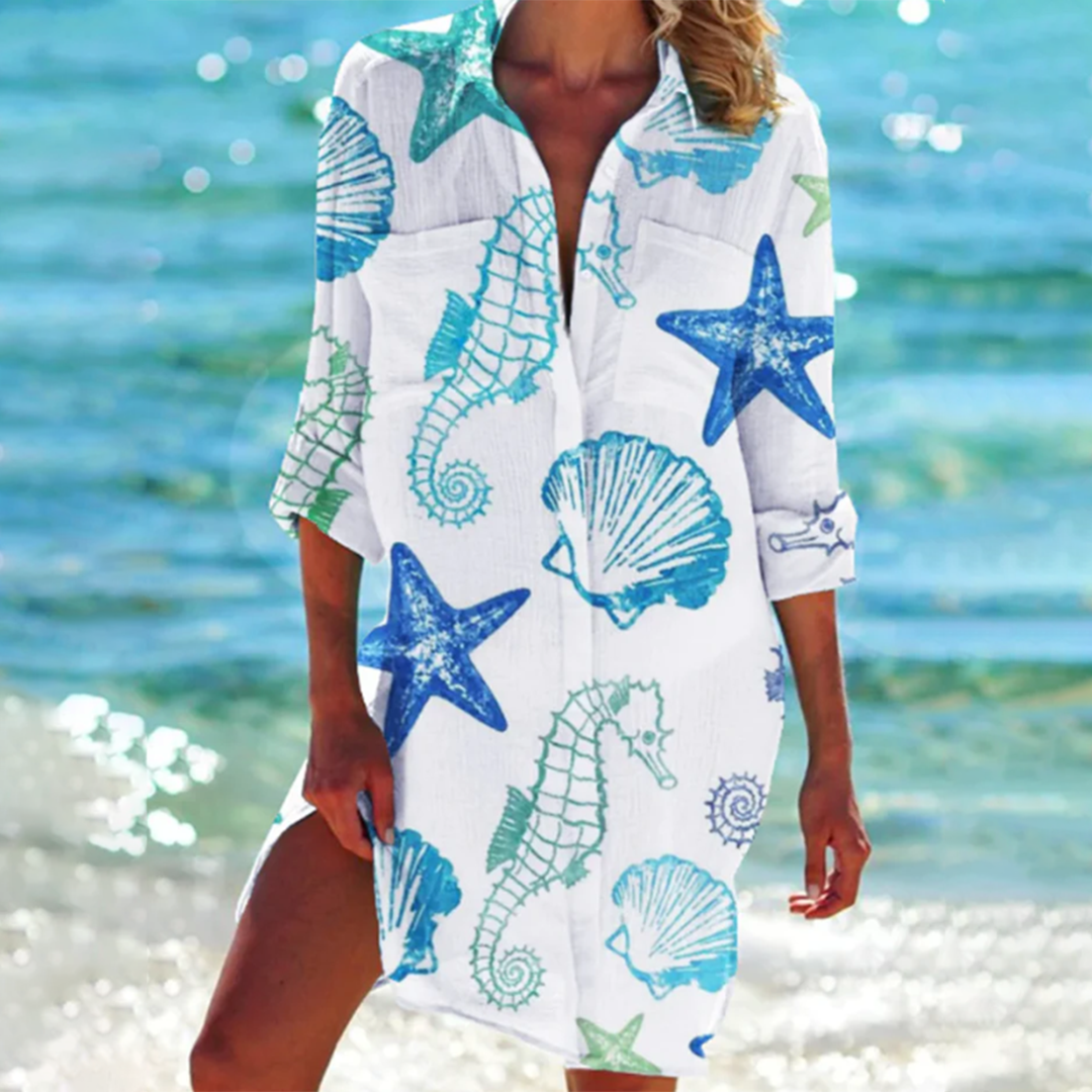Donna in spiaggia con copricostume bianco decorato con cavallucci marini, stelle marine e conchiglie blu e verdi, mare sullo sfondo.