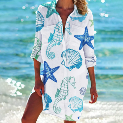 Donna in spiaggia con copricostume bianco decorato con cavallucci marini, stelle marine e conchiglie blu e verdi, mare sullo sfondo.