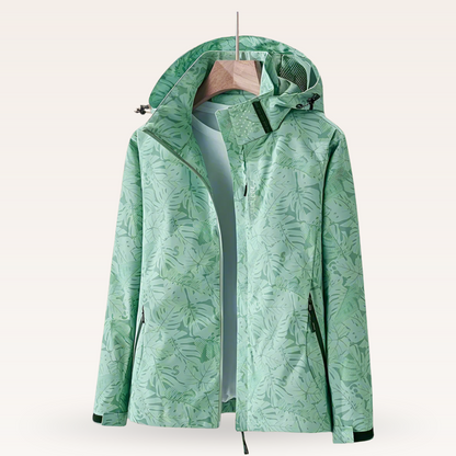 Giacca impermeabile verde con cappuccio, motivo foglie tropicali, appesa su gruccia. Abbigliamento outdoor, moda primavera, tessuto tecnico.