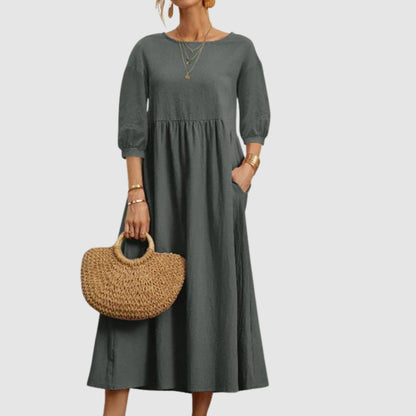 Abito lungo grigio con maniche a tre quarti, indossato con borsa di paglia e gioielli dorati. Moda donna elegante e casual.