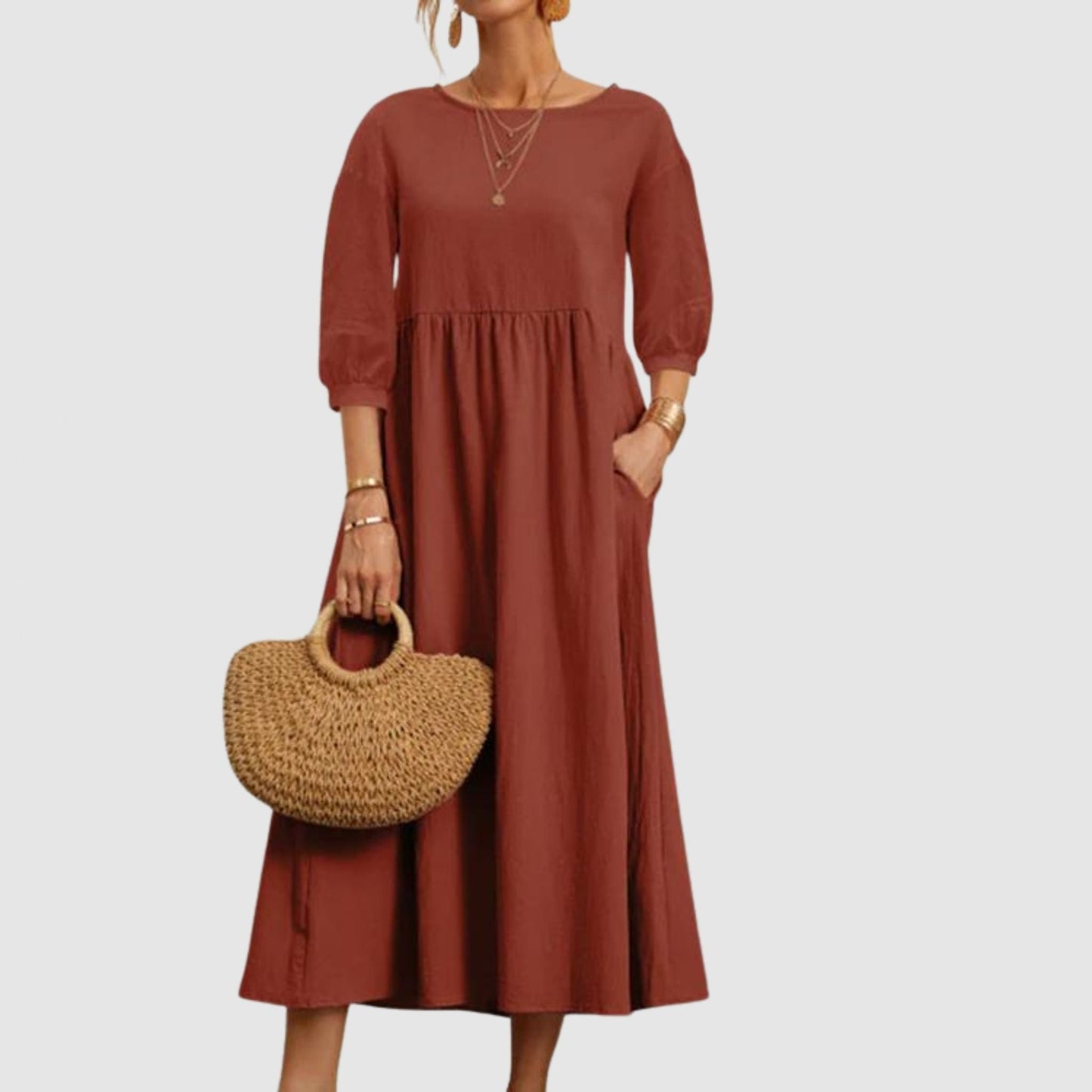 Abito lungo marrone con maniche a tre quarti, indossato da una donna con borsa di paglia. Moda femminile elegante e casual.