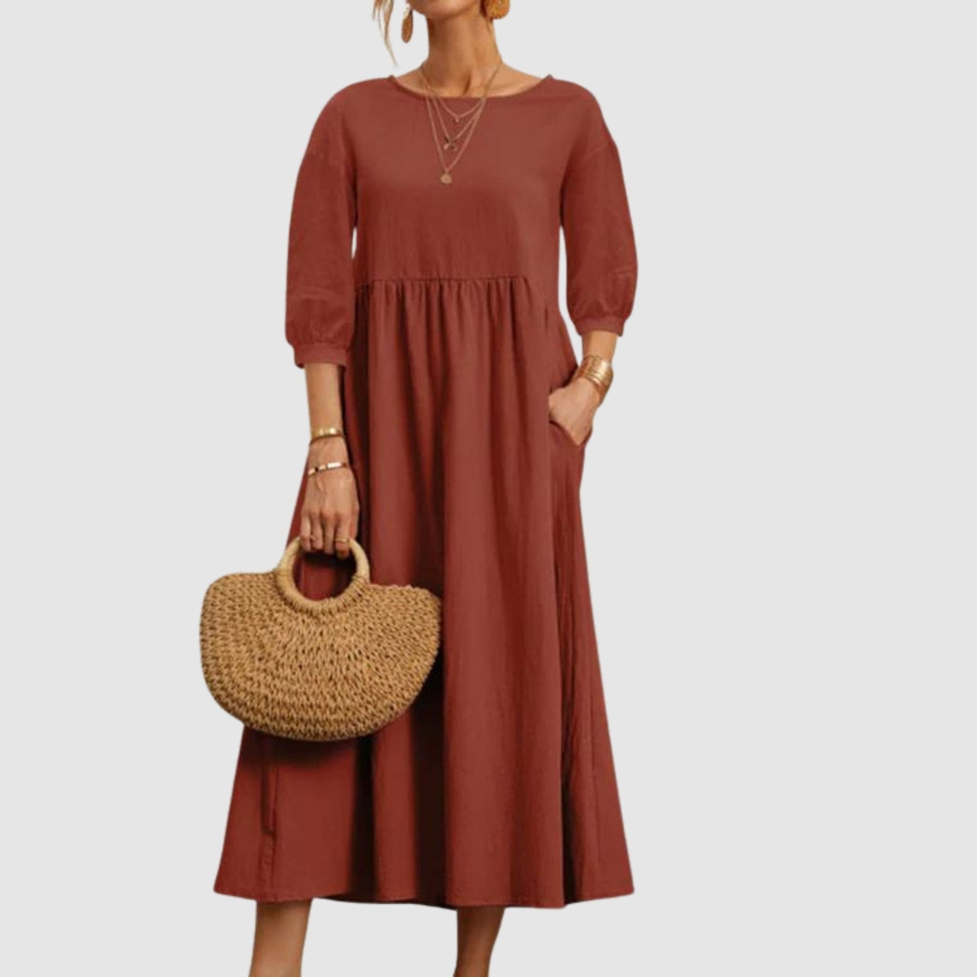 Abito lungo marrone con maniche a tre quarti, indossato da una donna con borsa di paglia. Moda femminile elegante e casual.