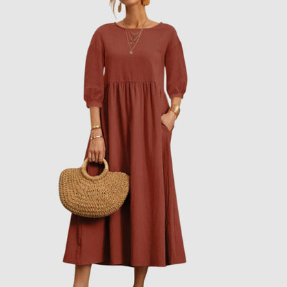 Abito lungo marrone con maniche a tre quarti, indossato da una donna con borsa di paglia. Moda femminile elegante e casual.