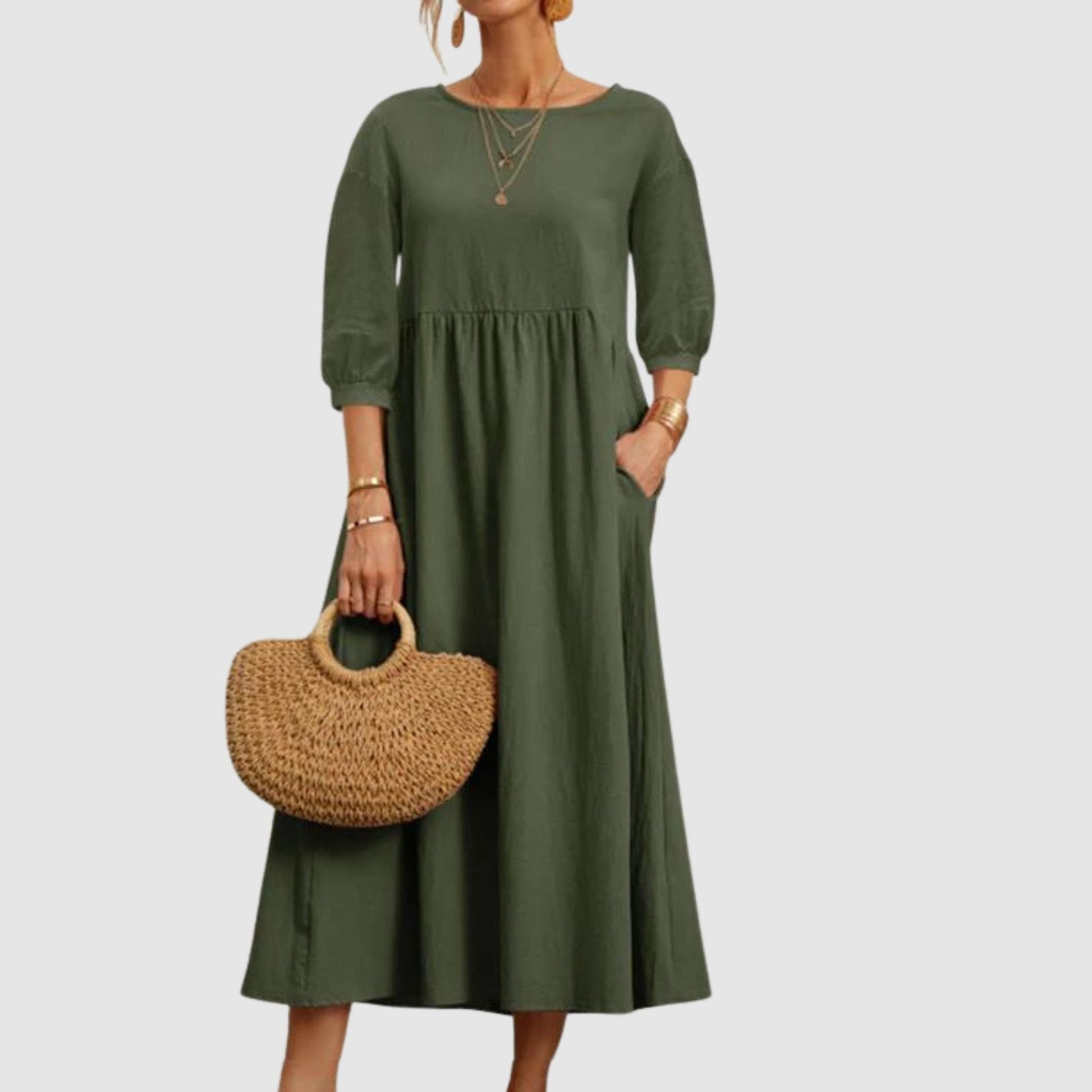 Abito lungo verde oliva con maniche a tre quarti, tasche laterali, accessori dorati e borsa in paglia. Moda donna elegante e casual.