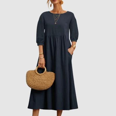Donna con abito lungo blu scuro, maniche a tre quarti, accessori dorati e borsa di paglia intrecciata. Moda elegante e casual.