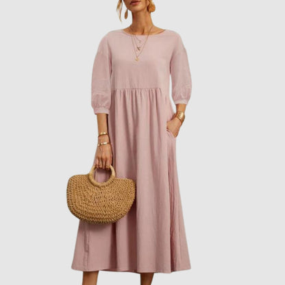 Abito lungo rosa donna con maniche a tre quarti, tasche laterali, accessori dorati e borsa paglia intrecciata. Moda femminile elegante.