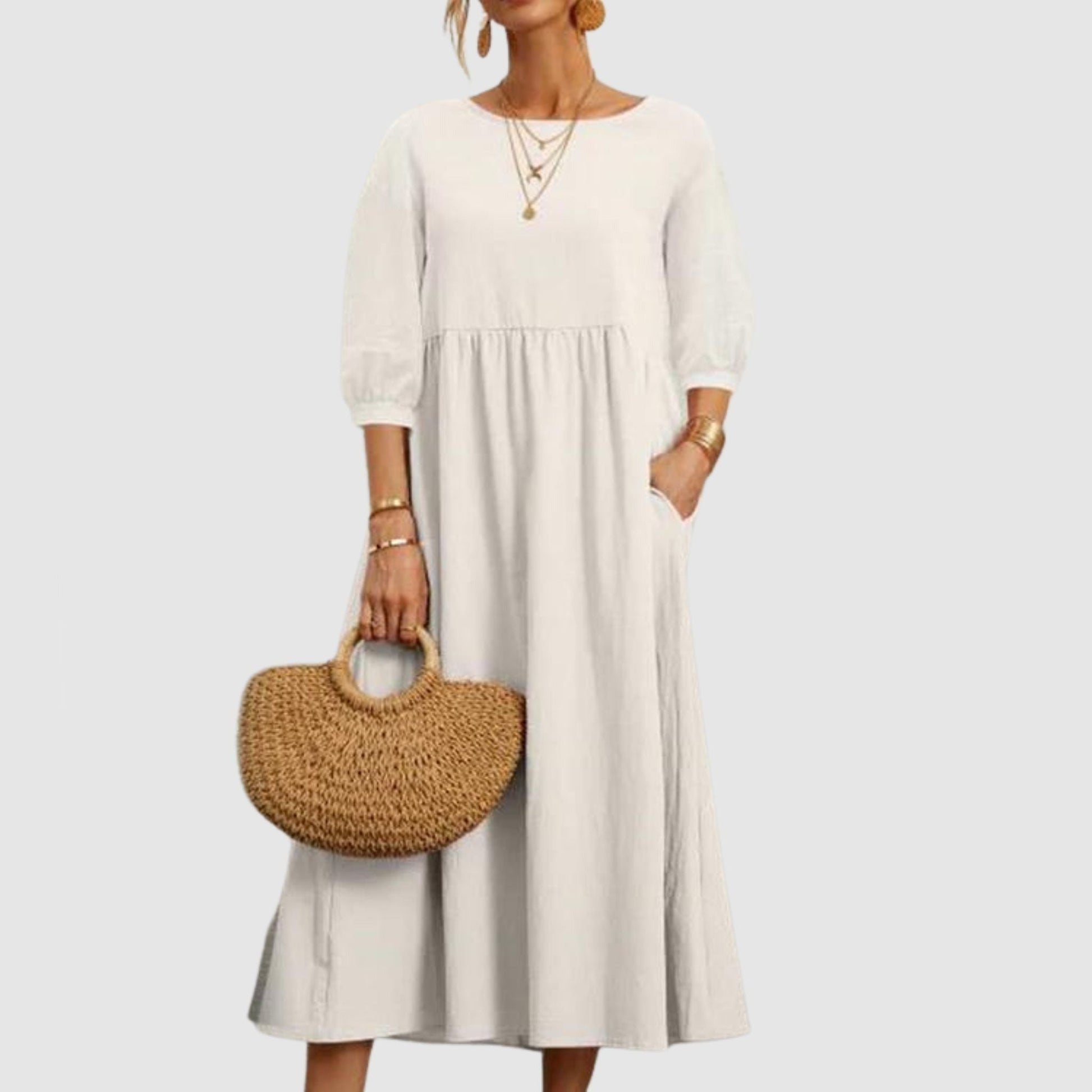 Abito lungo bianco con maniche a tre quarti, indossato con collane dorate e borsa in paglia. Moda donna elegante e casual.