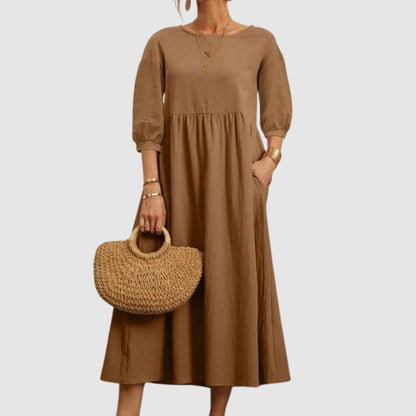 Abito lungo marrone con maniche a tre quarti, indossato con borsa in paglia intrecciata. Moda donna elegante, stile casual chic.