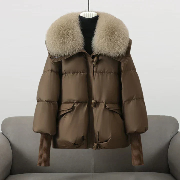 Giacca invernale marrone con cappuccio in pelliccia sintetica, design imbottito, maniche lunghe, su divano grigio. Moda donna, abbigliamento caldo.