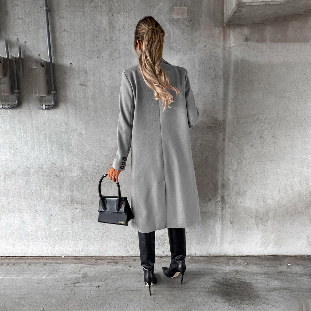Donna di spalle con cappotto grigio, stivali neri e borsa a mano nera, in un ambiente urbano con parete di cemento. Moda autunnale elegante.