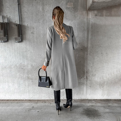 Donna di spalle con cappotto grigio, stivali neri e borsa a mano nera, in un ambiente urbano con parete di cemento. Moda autunnale elegante.