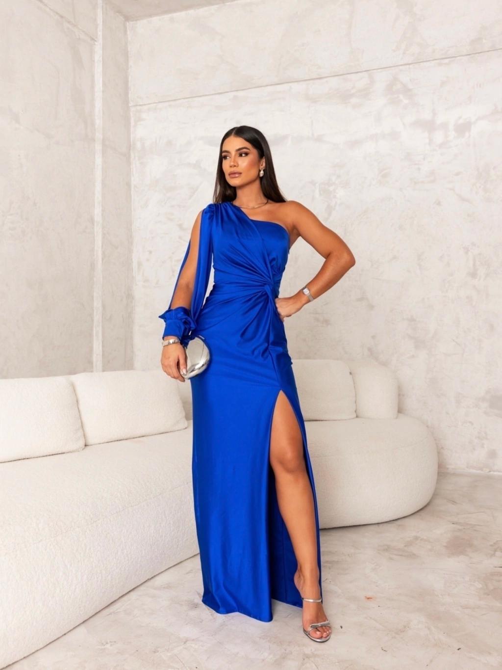 Donna in abito blu elegante con spacco alto, posa su divano bianco in stanza moderna. Moda femminile, abbigliamento da sera, stile glamour.