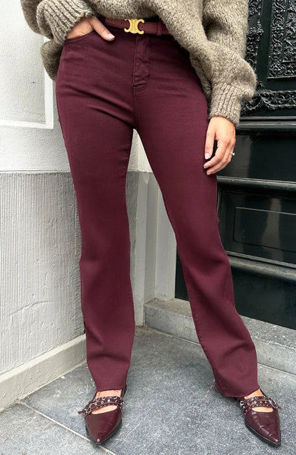 Donna con pantaloni bordeaux e maglione beige, scarpe a punta marroni con catena, posa su gradino. Moda autunnale, stile casual chic.