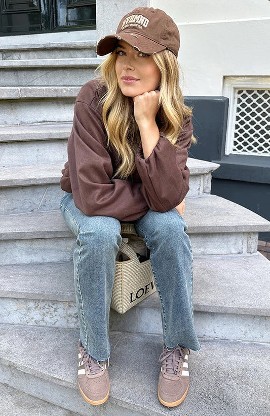 Donna seduta su gradini indossa cappello marrone, maglia marrone, jeans, scarpe sportive. Borsa di tela con scritta "LOEVE" accanto a lei. Moda casual urbana.