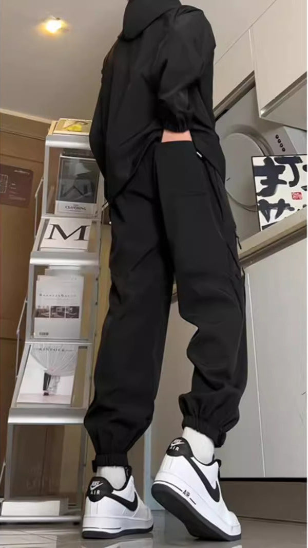 Persona in tuta nera e scarpe Nike Air Force 1 bianche e nere, in piedi accanto a scaffale con riviste. Look casual e moderno.