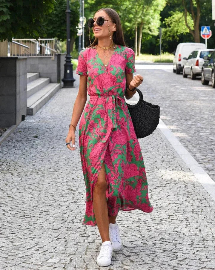 Donna con abito floreale rosa e verde, borsa nera intrecciata, occhiali da sole, cammina su strada acciottolata. Moda estiva, look casual chic.