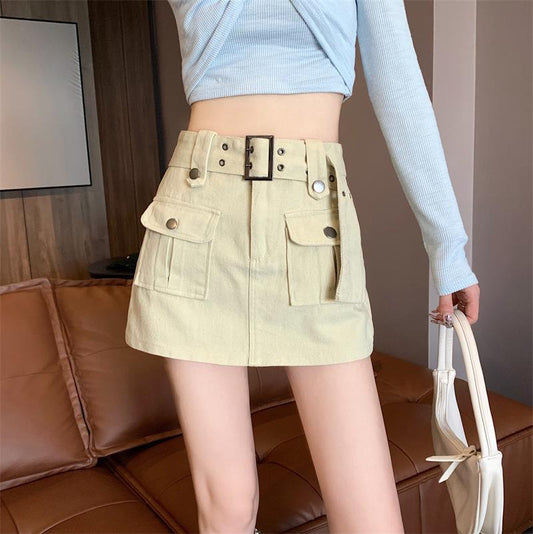 Minigonna beige con tasche e cintura, indossata da una persona con maglia azzurra, su divano marrone. Moda femminile, abbigliamento casual chic.