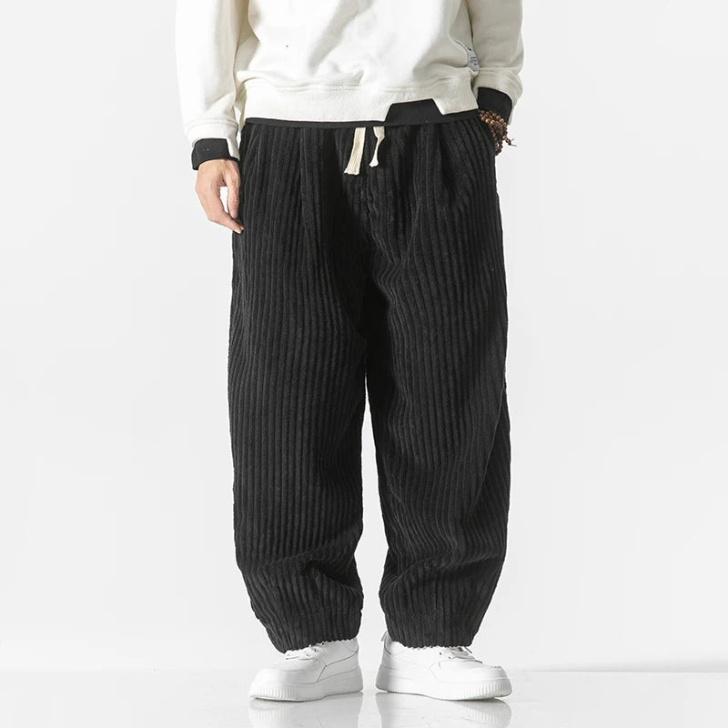 Pantaloni larghi a coste nere, indossati con felpa bianca e scarpe da ginnastica bianche. Stile casual, moda uomo, abbigliamento comodo.