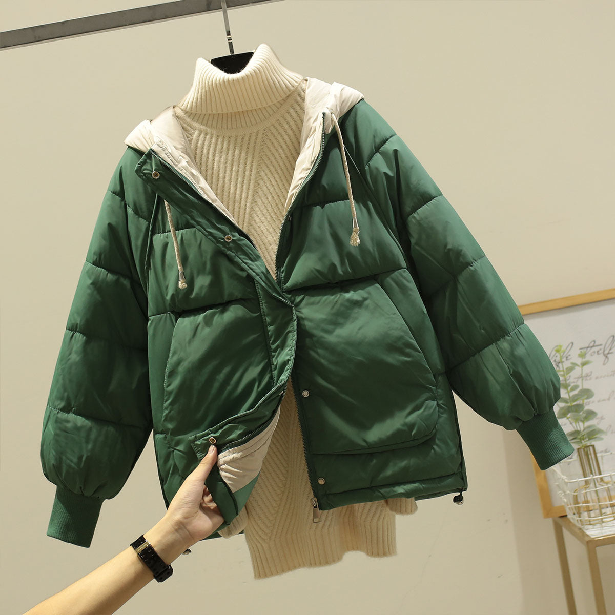 Giacca invernale verde imbottita con cappuccio, indossata sopra un maglione a collo alto color crema. Moda autunnale, abbigliamento caldo e casual.