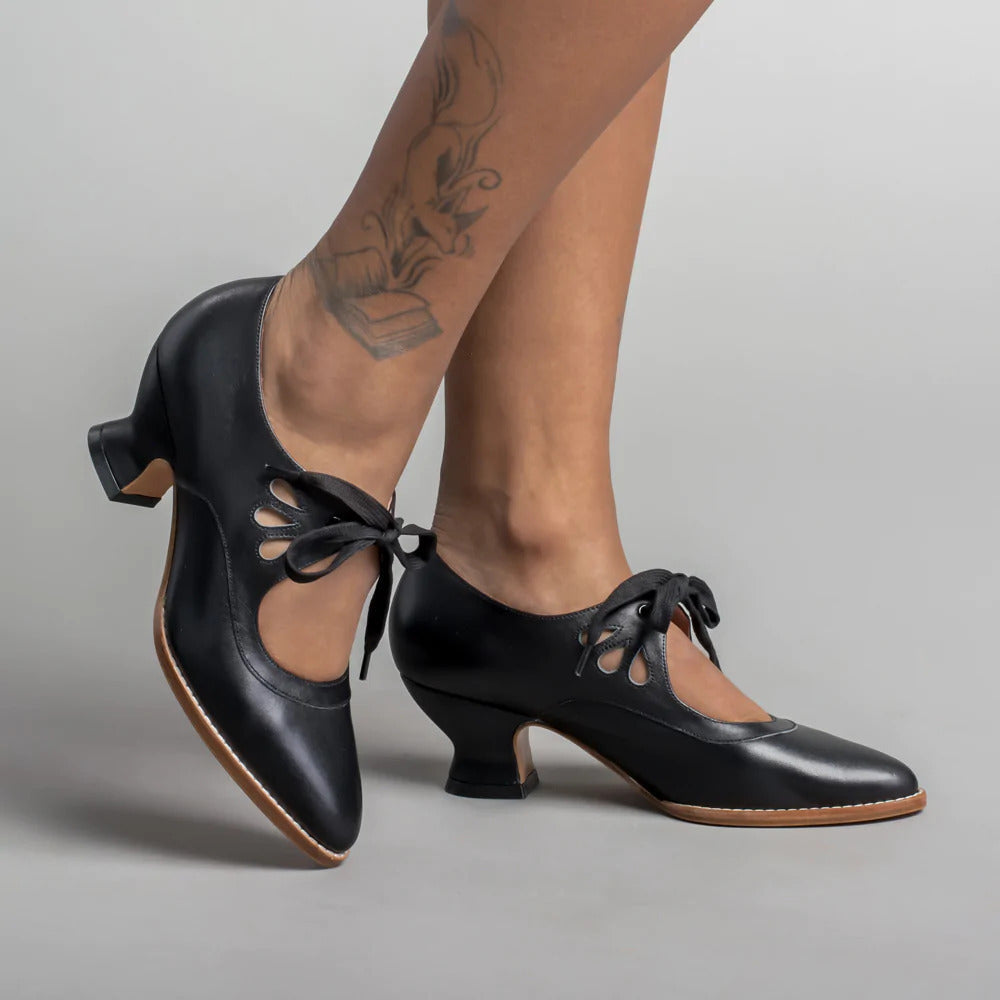 Scarpe da ballo nere con tacco basso, lacci e dettagli traforati, indossate su gambe con tatuaggio. Eleganti calzature per danza e spettacoli.
