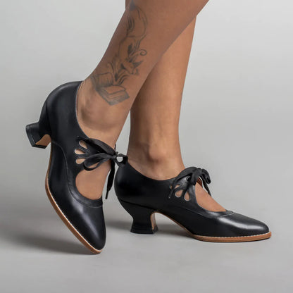 Scarpe da ballo nere con tacco basso, lacci e dettagli traforati, indossate su gambe con tatuaggio. Eleganti calzature per danza e spettacoli.