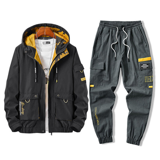 Completo sportivo uomo nero e grigio con giacca a vento e pantaloni cargo, dettagli gialli, cappuccio, tasche multiple, stile casual moderno.