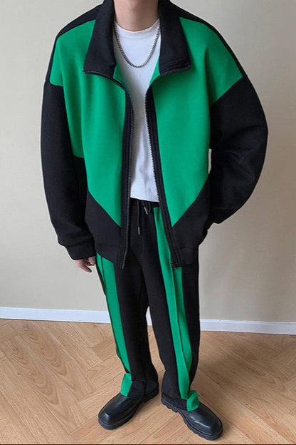 Uomo in tuta sportiva verde e nera, con maglietta bianca e collana, su pavimento in legno chiaro. Moda casual, abbigliamento sportivo trendy.