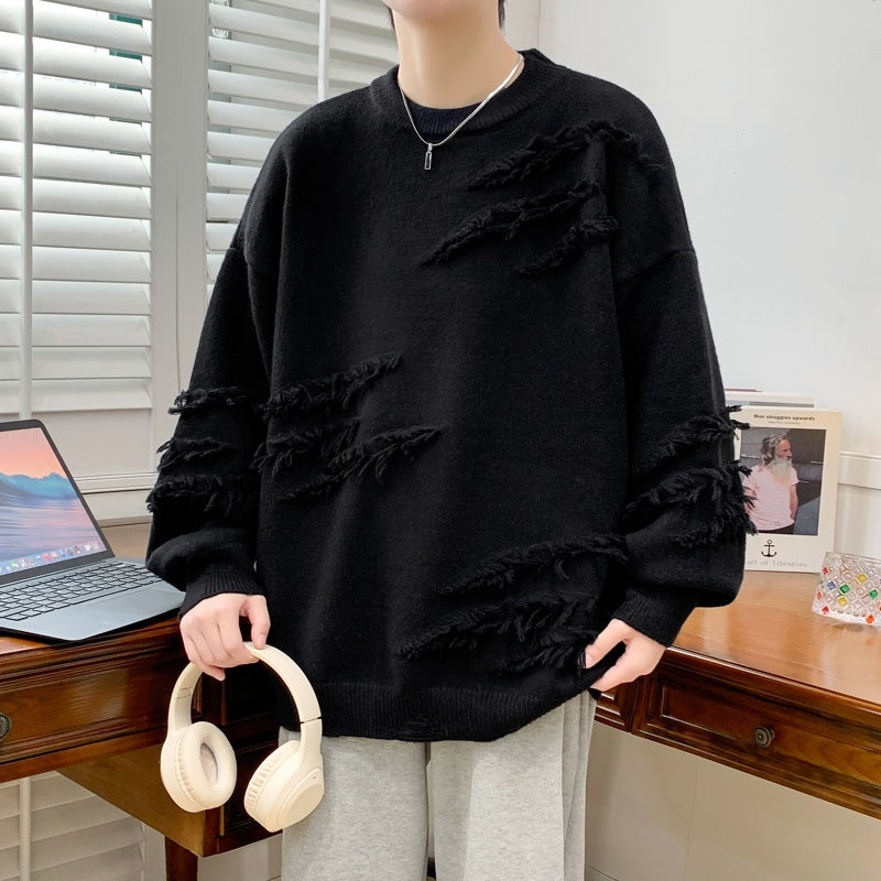 Maglione nero strappato, stile casual, indossato da persona in piedi accanto a laptop su scrivania di legno, cuffie bianche in mano.