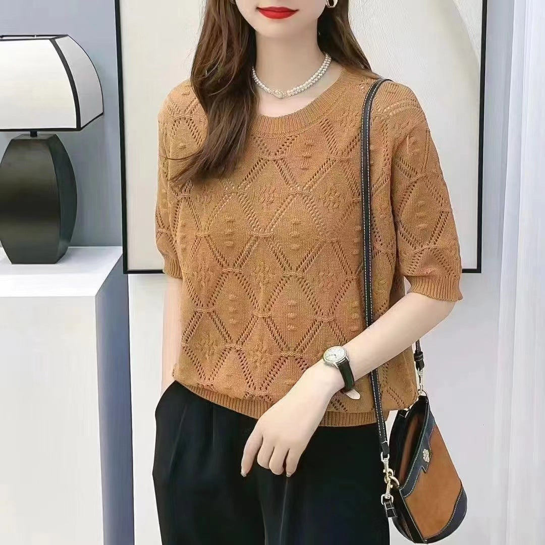 Donna con maglione marrone a maglia, borsa a tracolla nera e pantaloni neri. Indossa collana e orologio. Sfondo con lampada moderna.