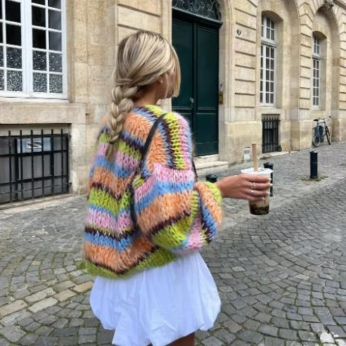 Donna con maglione colorato a righe e treccia, cammina su strada acciottolata con bevanda in mano. Sfondo di edifici storici. Moda autunnale urbana.