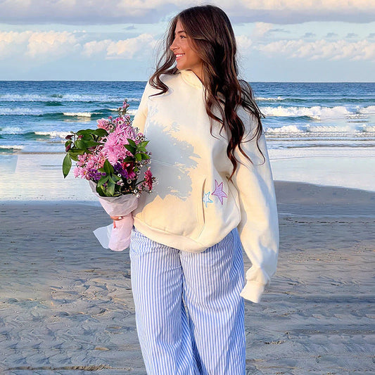 Donna sorridente in spiaggia con felpa bianca e pantaloni a righe, tiene un mazzo di fiori rosa. Mare e cielo nuvoloso sullo sfondo.