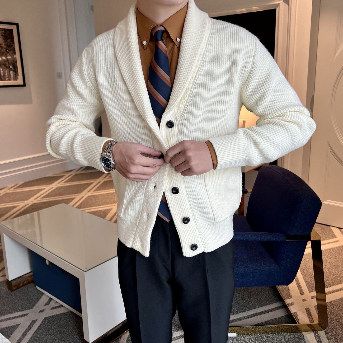 Uomo in cardigan bianco con cravatta a righe, camicia marrone e pantaloni neri in un salotto elegante. Moda maschile, abbigliamento autunnale.