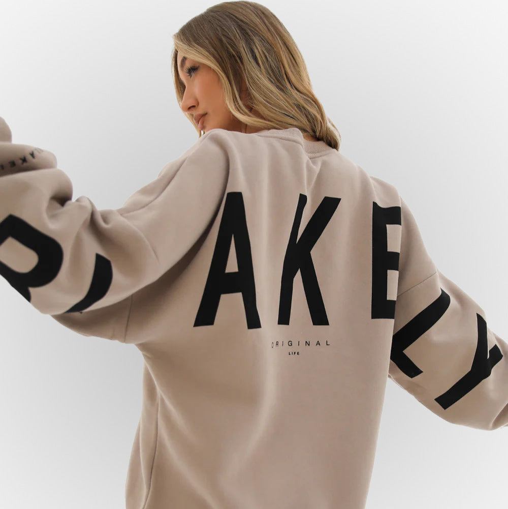 Donna con felpa beige oversize, scritta "AKELY" in nero. Moda casual, abbigliamento streetwear, stile urbano, tendenze autunno 2023.