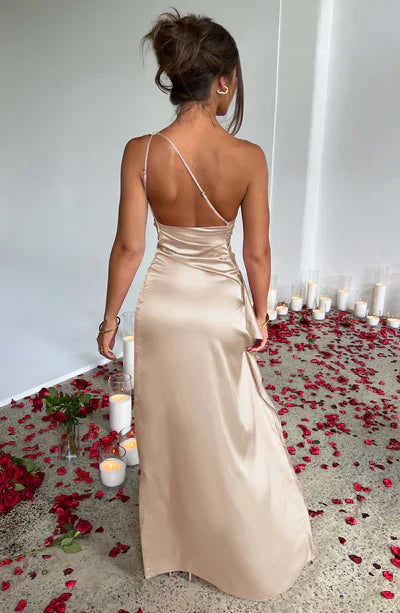 Donna in abito lungo di seta beige, schiena scoperta, circondata da petali di rosa rossa e candele accese su pavimento grigio. Eleganza e romanticismo.