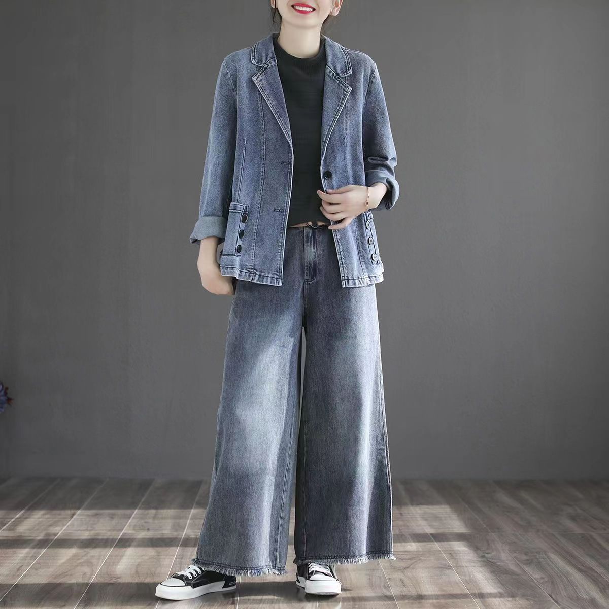 Donna in giacca e pantaloni di jeans larghi, stile casual, su sfondo grigio. Moda denim, abbigliamento femminile trendy, scarpe da ginnastica nere.