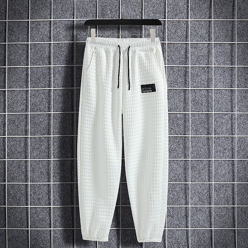 Pantaloni sportivi bianchi a quadri, con elastico in vita e coulisse nera, appesi su griglia metallica. Stile casual, moda uomo.